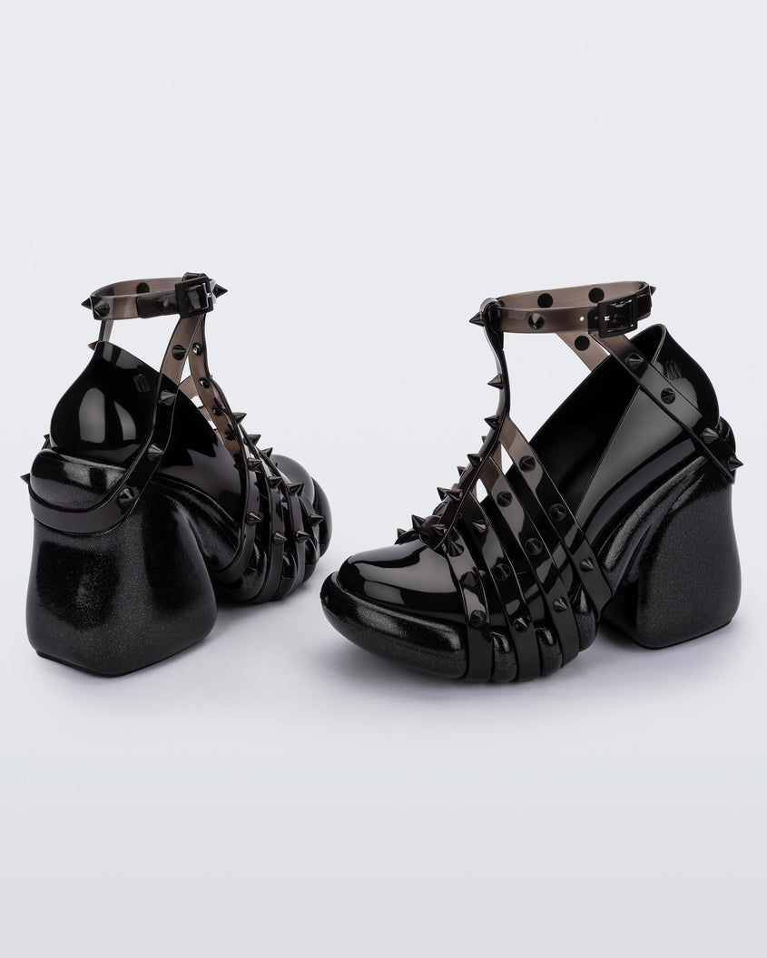 An angled side and back view of a pair of black/clear Melissa Punk Love Heels with black straps fastening at a top ankle strap with spike details.