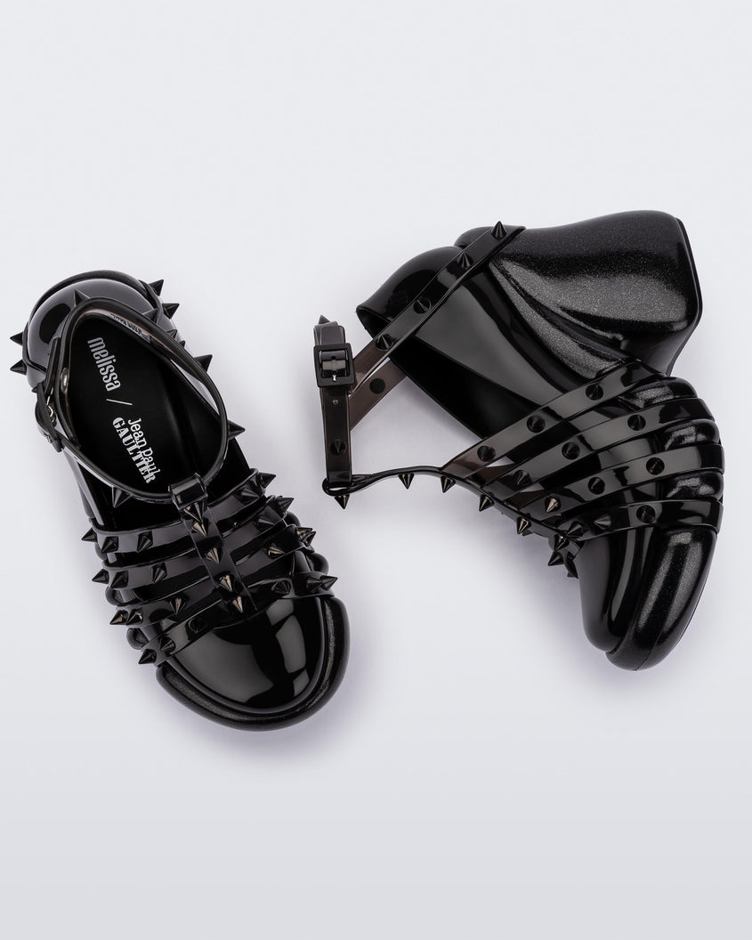A top and side view of a pair of black/clear Melissa Punk Love Heels with black straps fastening at a top ankle strap with spike details.