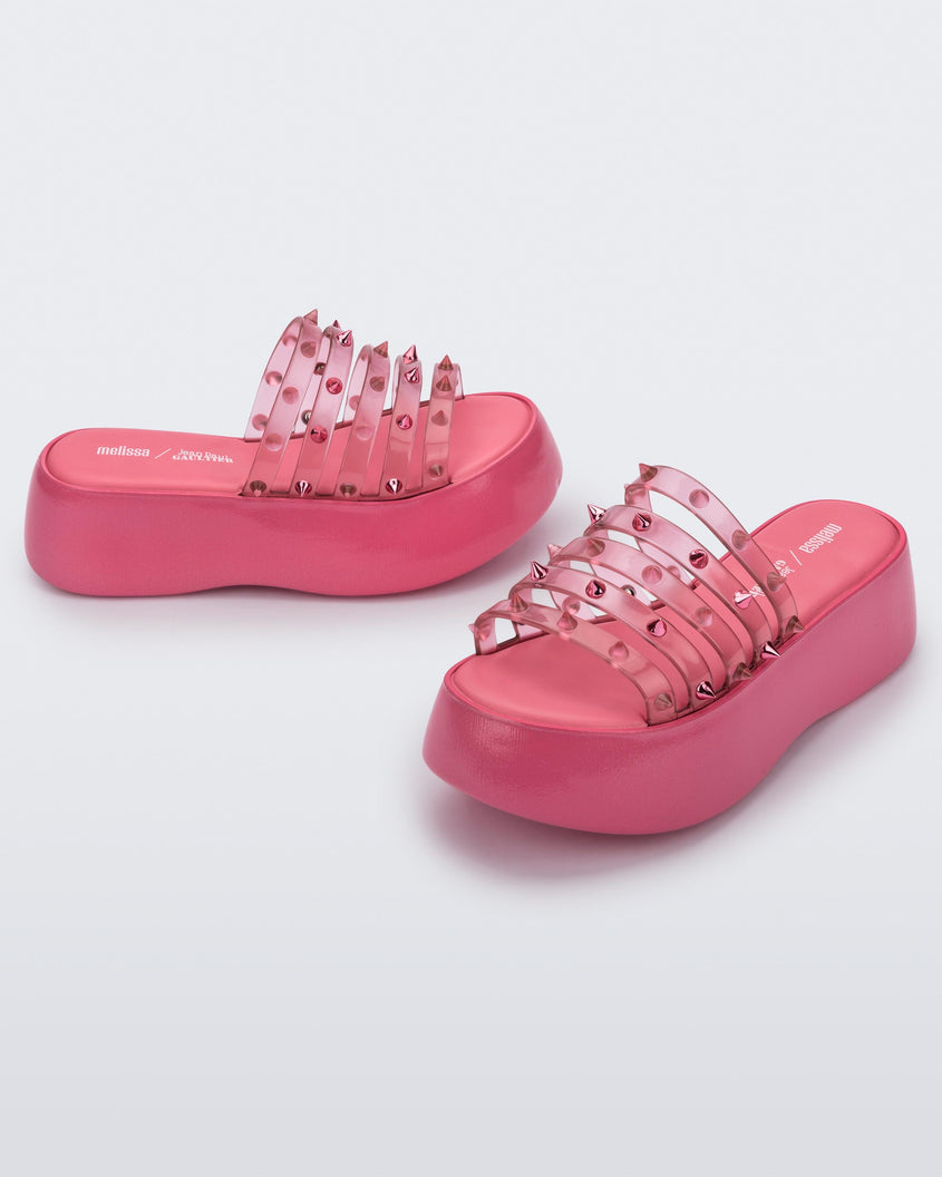 An angled front and side view of a pair of Pink/Transparent Pink Melissa Punk Love Becky platform slides with a pink base and several top straps with spike stud details.