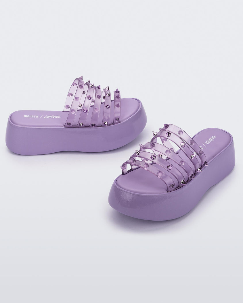 An angled front and side view of a pair of violet / clear lilac Melissa Punk Love Becky platform slides with a lilac base and several top straps with spike stud details.