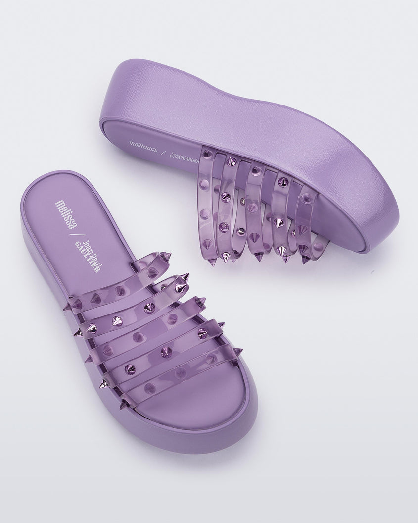 A top and side view of a pair of violet / clear lilac Melissa Punk Love Becky platform slides with a lilac base and several top straps with spike stud details.