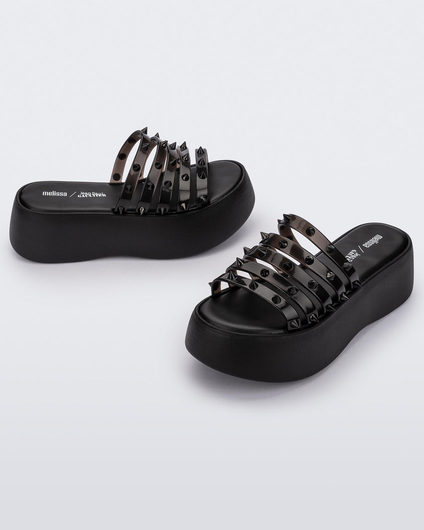 An angled front and side view of a pair of Black/Clear Black Melissa Punk Love Becky platform slides with a black base and several top straps with spike stud details.