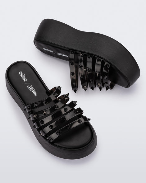 A top and side view of a pair of Black/Clear Black Melissa Punk Love Becky platform slides with a black base and several top straps with spike stud details.