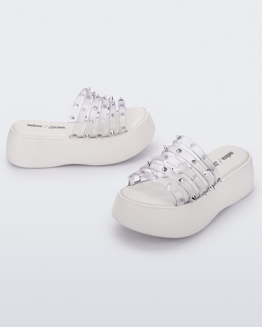 An angled front and side view of a pair of White/Clear Melissa Punk Love Becky platform slides with a white base and several top straps with spike stud details.