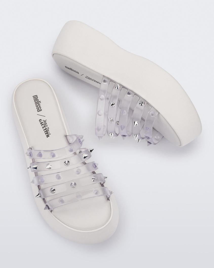 A top and side view of a pair of White/Clear Melissa Punk Love Becky platform slides with a white base and several top straps with spike stud details.