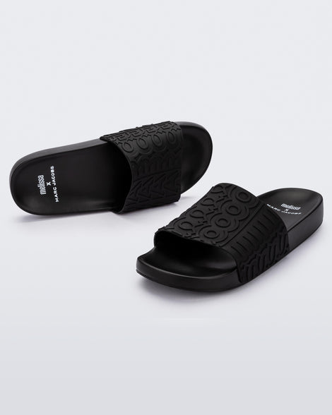 An angled front and top view of a pair of black Melissa Slides with the Marc Jacobs logo across the front strap.