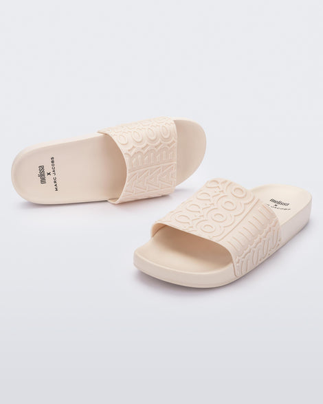 An angled front and top view of a pair of beige Melissa Slides with the Marc Jacob's logo across the front strap.