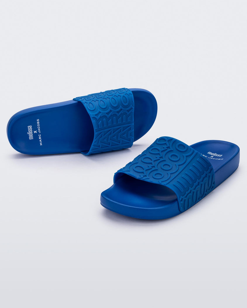 An angled front and top view of a pair of blue Melissa Slides with the marc jacob's logo across the front strap.