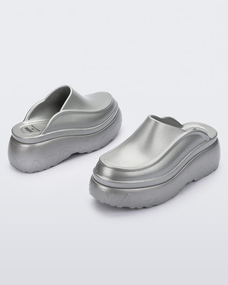 An angled front and back view of a pair of silver Melissa Clogs with the Marc Jacob's logo across the sole.