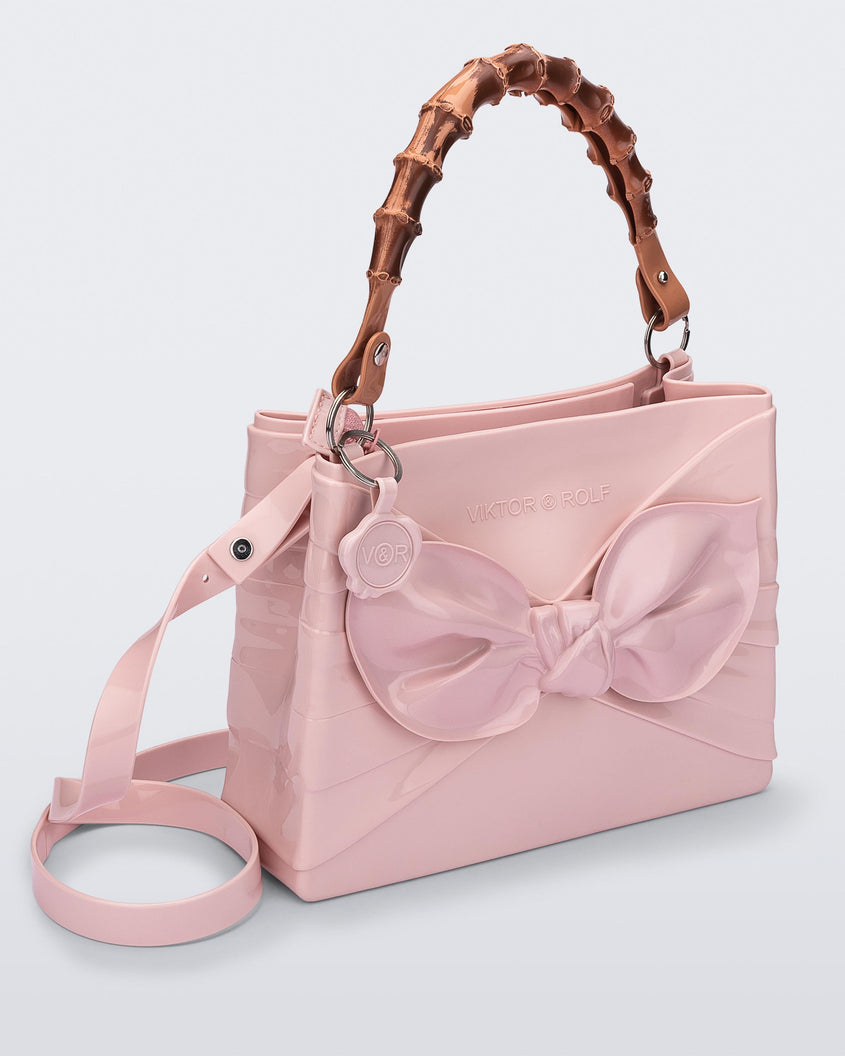 Angled view of the Melissa Tie handag in pink with long shoulder strap, 3D bow detail on the front and natural colored bamboo shaped top handle.