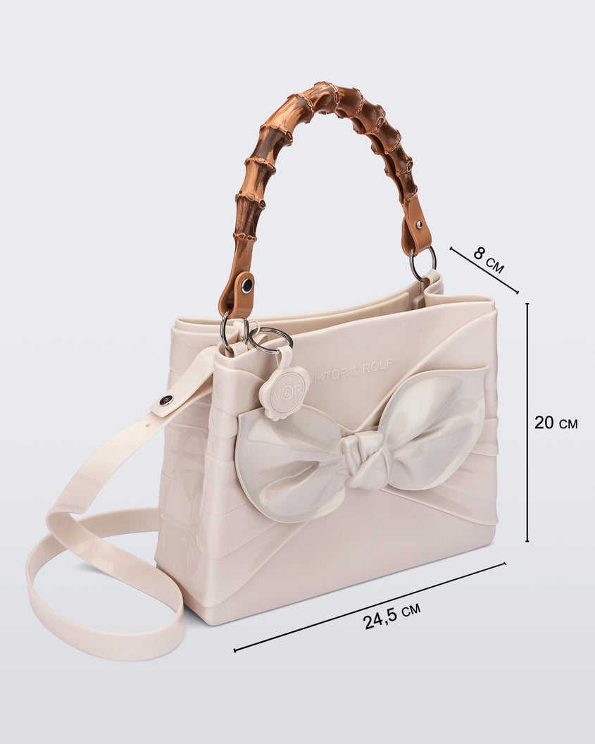 An angled side view of a beige Melissa Tie Bag with a bow detail on the front, two bamboo top straps and a beige shoulder strap. The image also shows the dimensions of the bag which reads 24.5 centermeters wide, 20 centermeters high and 8 centermeters deep.