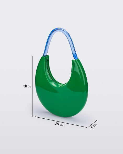 An angled front view of a Green/Blue Melissa Moon Bag with a crescent shaped green bag base and a blue beige top strap. The image also shows the dimensions of the bag which reads 30 centermeters high, 29 centermeters wide and 6 centermeters deep.