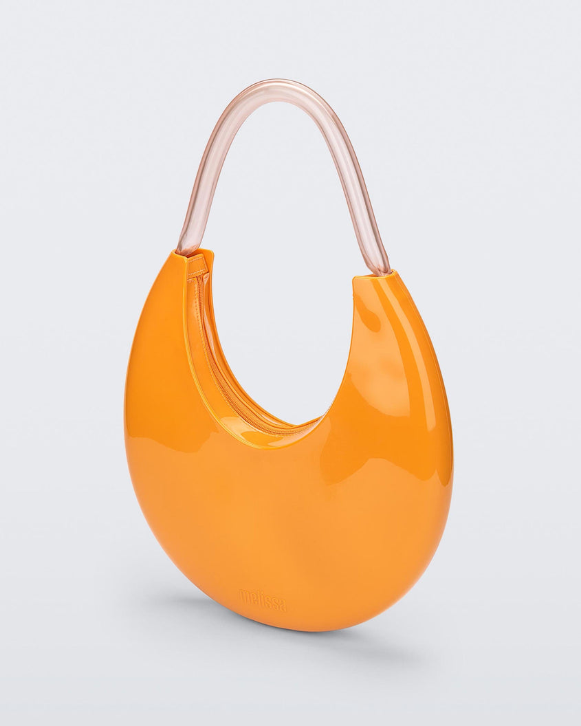 An angled front view of a orange/beige Melissa Moon Bag with a crescent shaped orange bag base and a clear beige top strap.