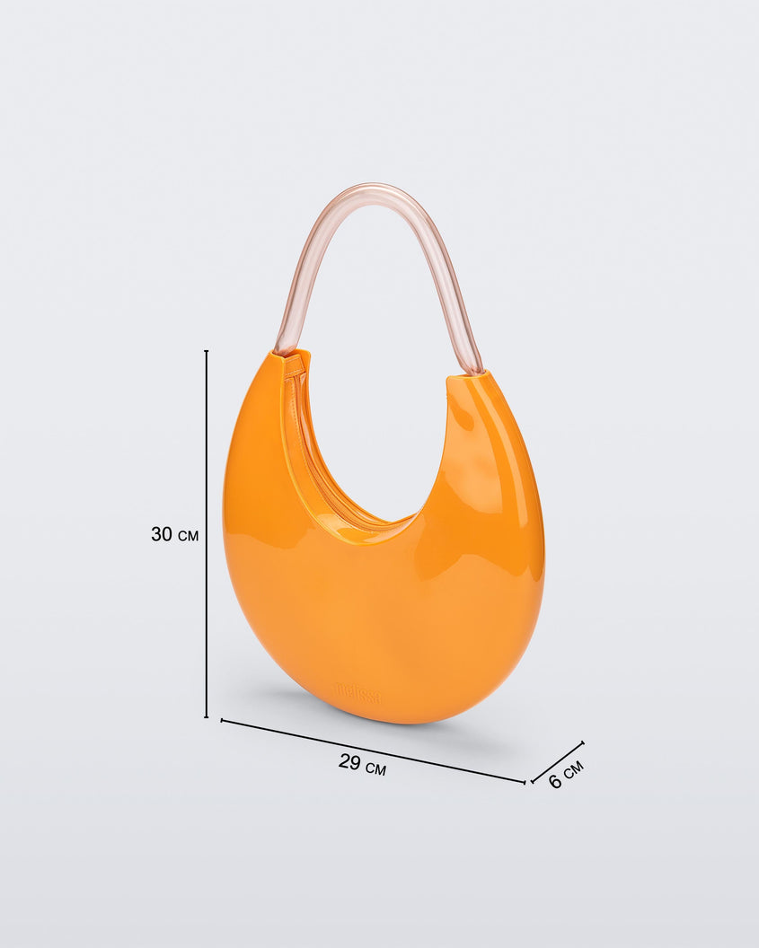 An angled front view of a orange/beige Melissa Moon Bag with a crescent shaped orange bag base and a clear beige top strap. The photo also shows the dimensions of the bag which read 30 centermeters high, 29 centermeters wide and 6 centermeters deep.