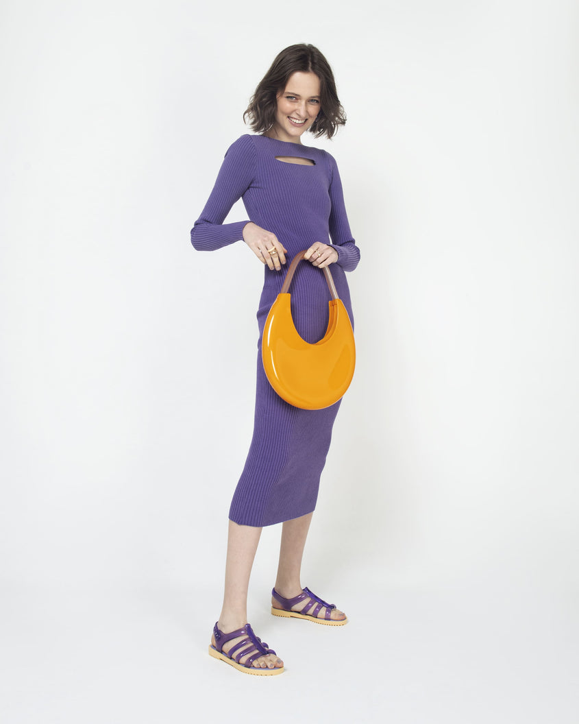 A model posing for a picture in a purple dress, and showing an orange/beige Melissa Moon Bag with a crescent shaped orange bag base and a clear beige top strap.