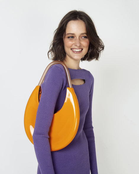 A zoomed in shot of a model posing for a picture in a purple dress, and showing an orange/beige Melissa Moon Bag with a crescent shaped orange bag base and a clear beige top strap on her shoulder.