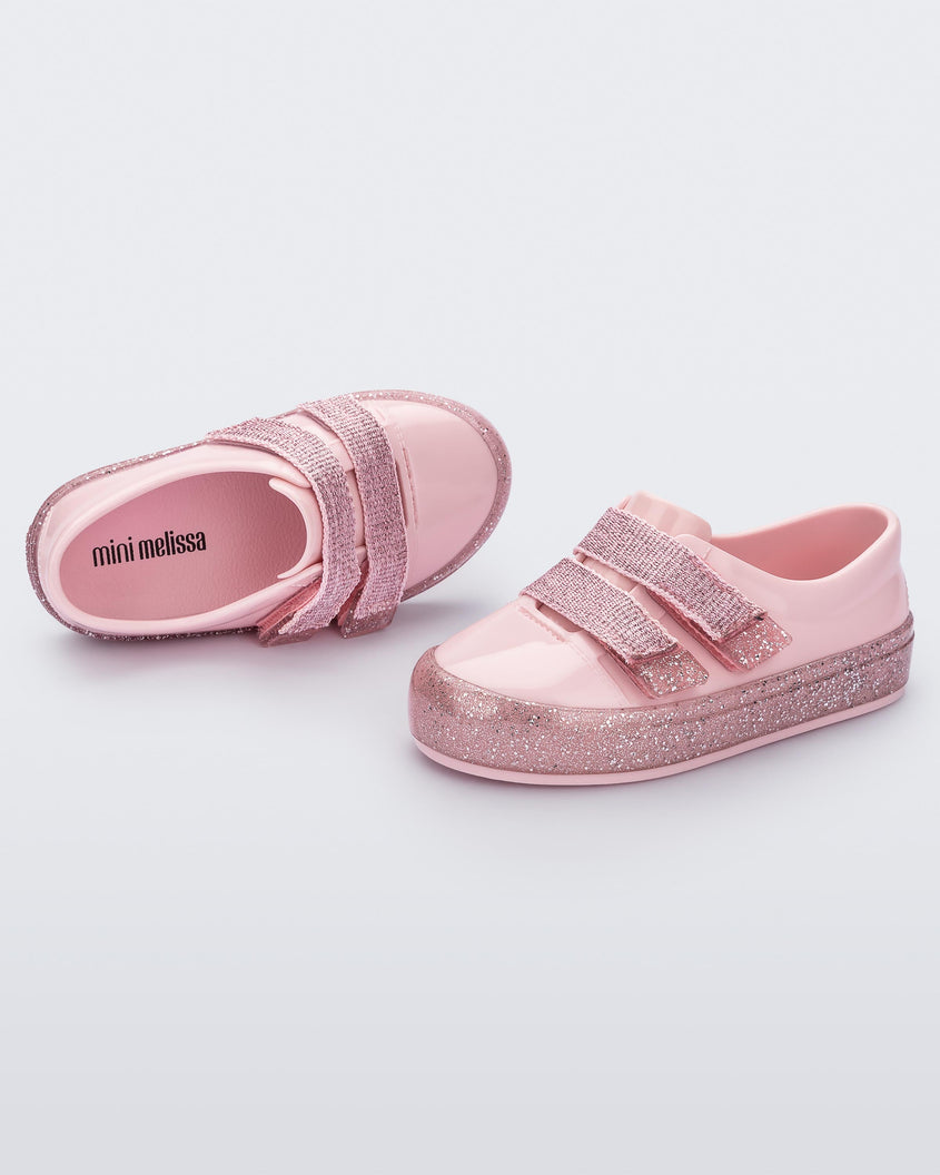 An angled side and top view of a pair of Pink/Pink Glitter Mini Melissa Beanny Bugs sneakers with a pink base, two shiny pink velcro straps and a pink glitter sole.