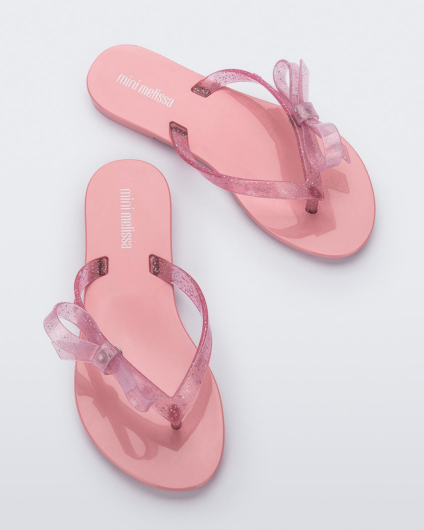 Top view of a pair of Pink/Glitter Pink Mini Melissa Harmonic Sweet flip flops with a pink sole, pink glitter straps with a bow on top.