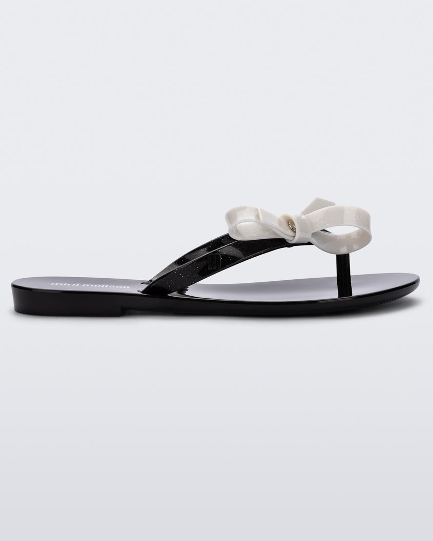 Side view of a black/glitter white Mini Melissa Harmonic Sweet flip flop with a black base, black glitter straps and a white bow on top.