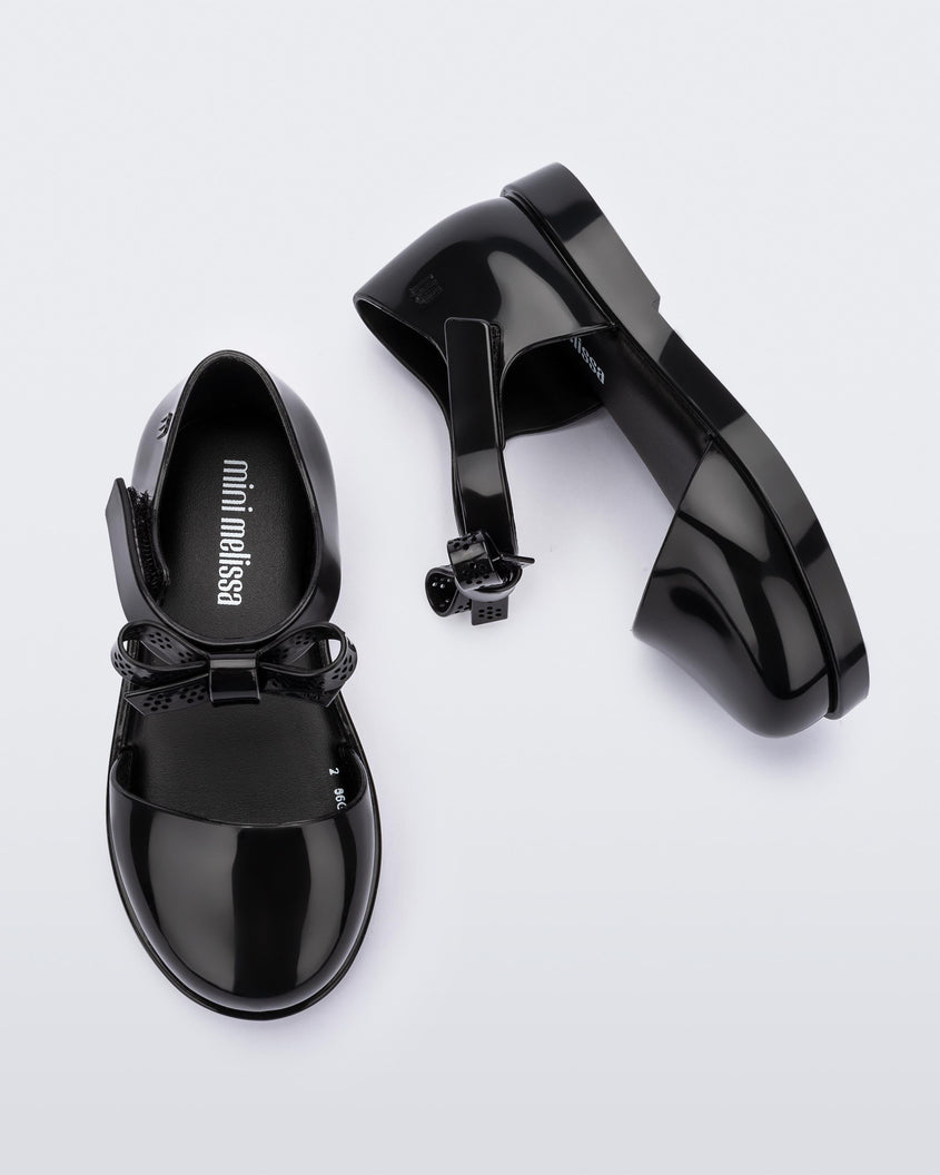 A top and side view of a pair of Black Mini Melissa Amy sandals with a covered toe section and a single black strap with a lace like bow detail.