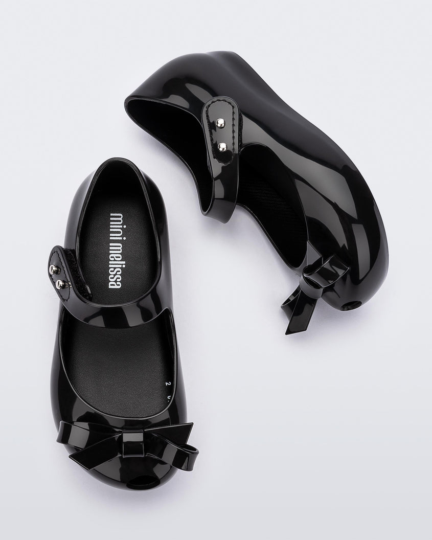 Top and side view of a pair of black Mini Melissa Ultragirl Bow flats with a black base, top strap and black bow detail on the top.