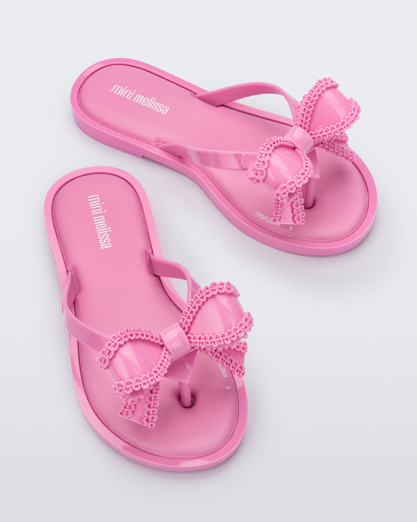 A front and top view of a pair of pink Mini Melissa Slim flip flops with a lace like bow detail on the front strap.
