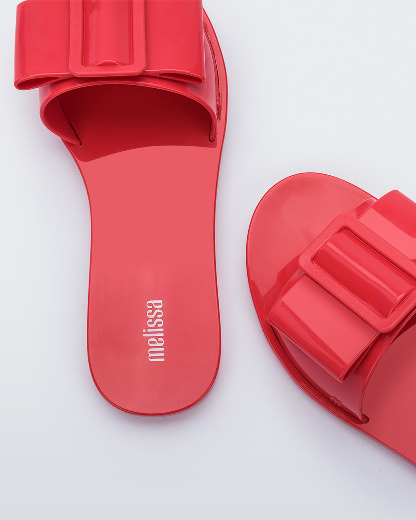 A zoomed in view of a pair of red Melissa Babe slides with a buckle like bow detail on the front strap, showing the melissa logo on the insole and an m logo on the outter strap.