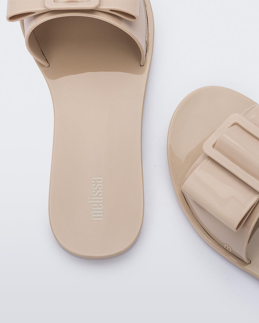 A zoomed in view of a pair of Beige Melissa Babe slides with a buckle like bow detail on the front strap, showing the melissa logo on the insole and an m logo on the outter strap.