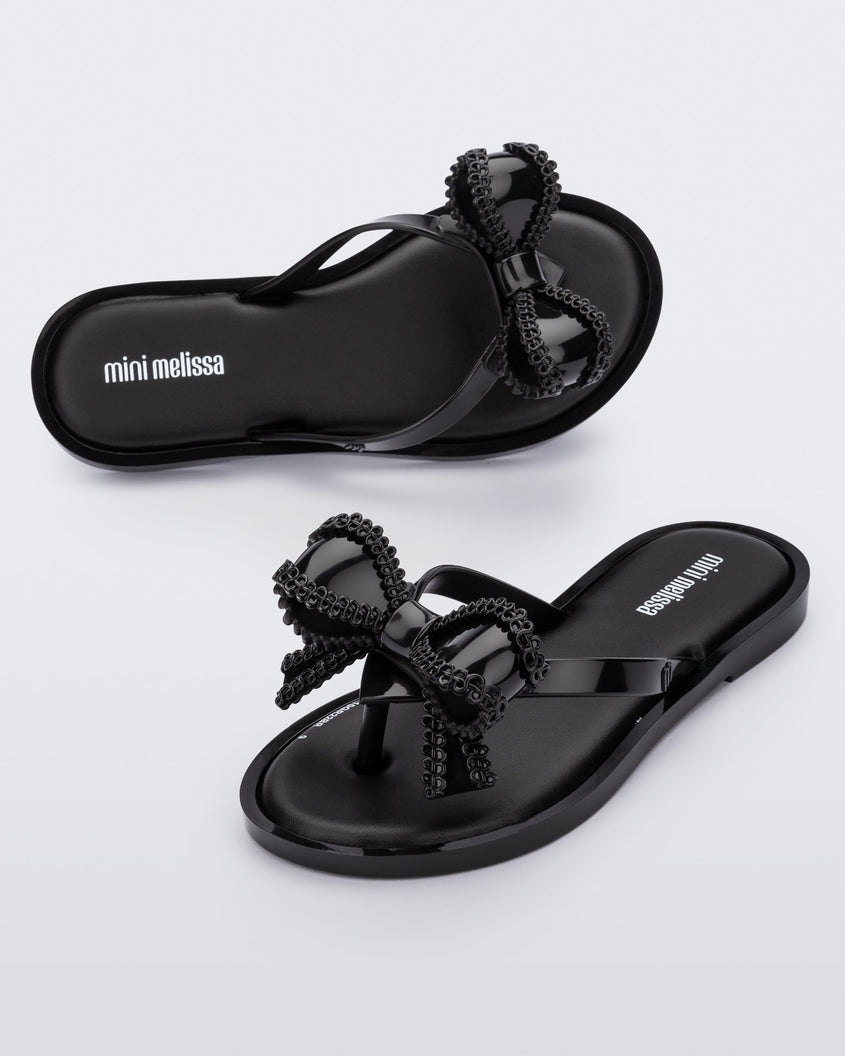 An angled side and top view of a pair of Black Mini Melissa Slim flip flops with a lace like bow detail on the front straps.