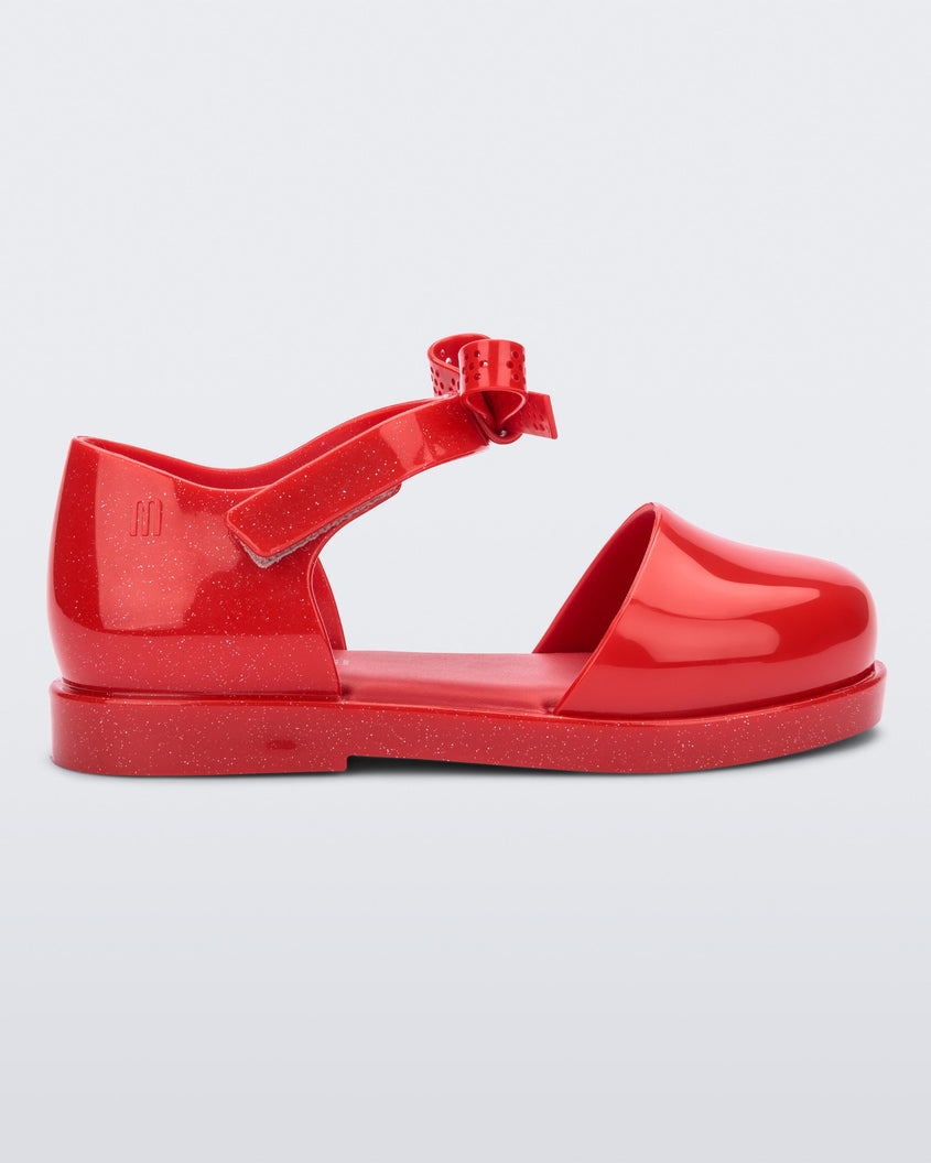 Side view of a red Mini Melissa Amy sandal with a red closed toe section and a back red glitter strap with a lace like bow detail.