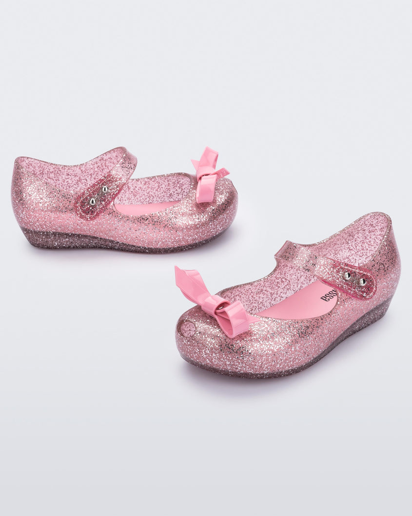 An angled front and side view of a pair of Glitter Pink Mini Melissa Ultragirl Bow flats with a pink glitter base, top strap and pink bow on the toe.