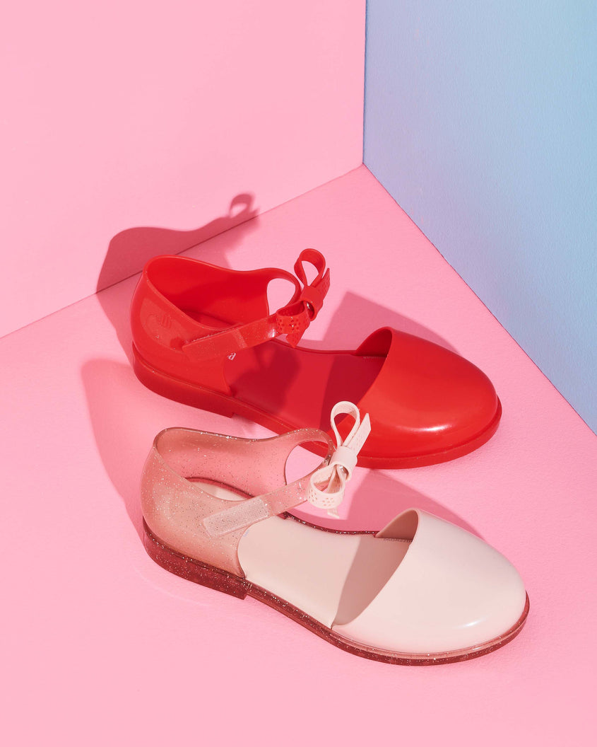 An angled top and side view of a red Mini Melissa Amy sandal and a pink Mini Melissa Amy sandal both with a closed toe section and a back strap with a lace like bow detail.