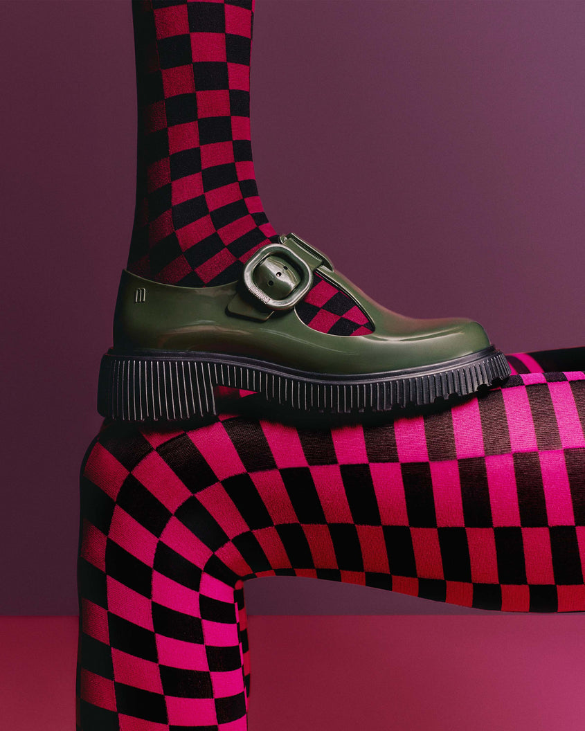 A side view of a model's leg in pink and black checkered tights, wearing a pair of Green Mini Melissa Jackie loafer with a green base, two cut outs, a green buckle detail strap and black sole, standing on another model's legs in the same tights, 
