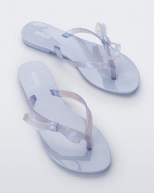 An angled top view of a pair of Lilac/Pearly blue Melissa Harmonic Sweet flip flops with a lilac/pearly blue sole, straps and bow on top.