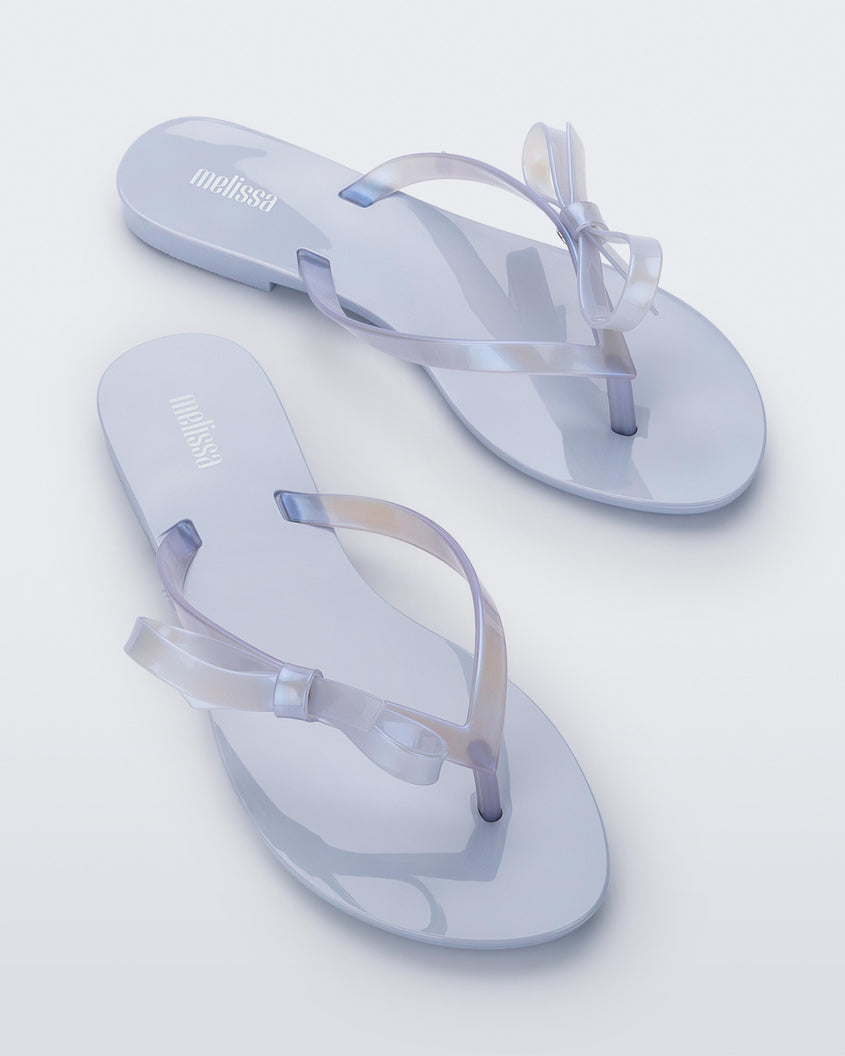 An angled top view of a pair of Lilac/Pearly blue Melissa Harmonic Sweet flip flops with a lilac/pearly blue sole, straps and bow on top.