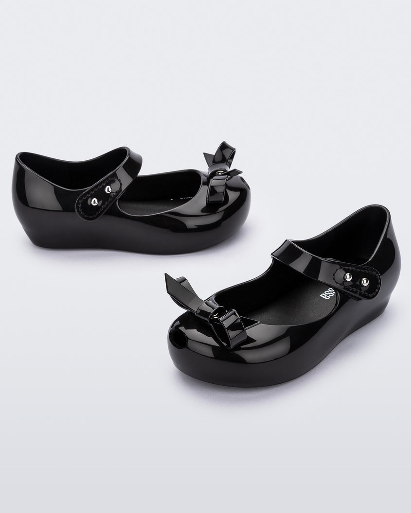 Side view of a pair of black Mini Melissa Ultragirl Bow flats with a black base, top strap and black bow detail on the top.