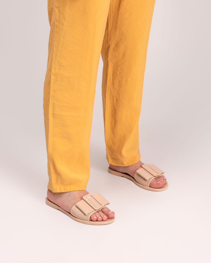 A model's legs wearing yellow pants and a pair of Beige Melissa Babe slides with a buckle like bow detail on the front strap.