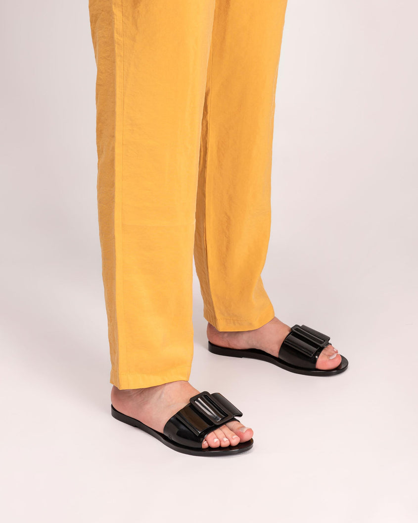 A model's legs wearing yellow pants and a pair black Melissa Babe slide with a buckle like bow detail on the front strap.