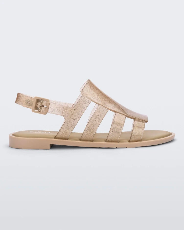 Side view of a transparent glitter beige Melissa Boemia sandal with straps conjoining in the front and a back buckle.