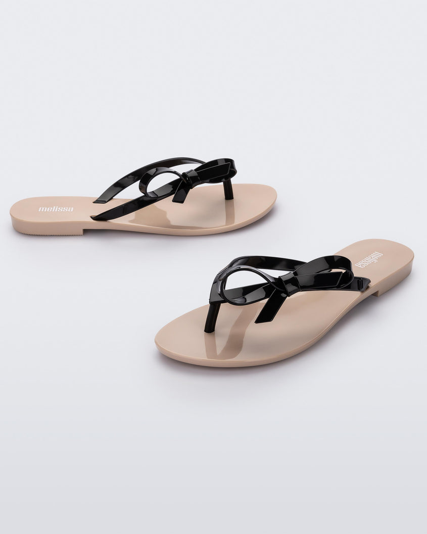An angled front and side view of a pair of beige/black Melissa Harmonic Sweet flip flops with a beige sole and black straps with a bow detail on the front.