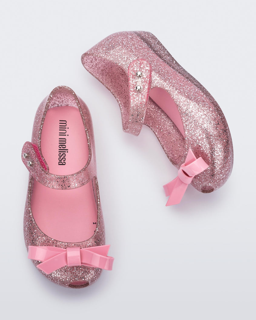 A top and side view of a pair of Glitter Pink Mini Melissa Ultragirl Bow flats with a pink glitter base, top strap and pink bow on the toe.
