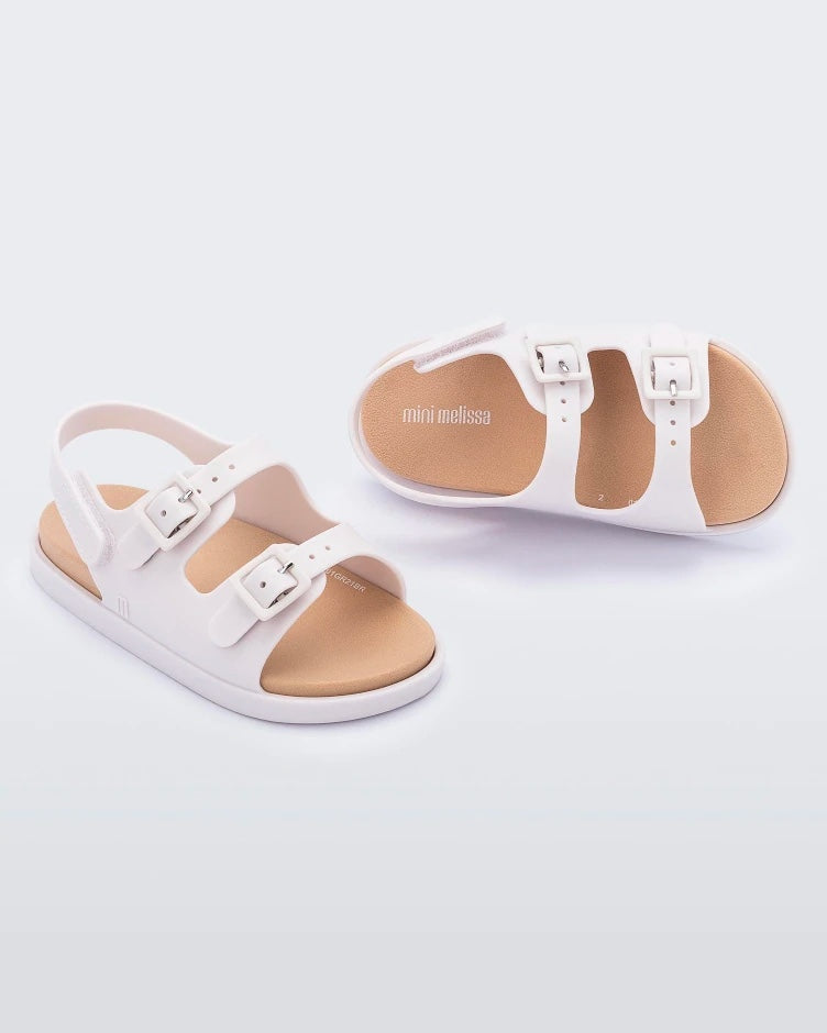Angled side and top view of a pair of beige Mini Melissa Wide Sandals with a beige base, two buckles on top and an ankle strap.