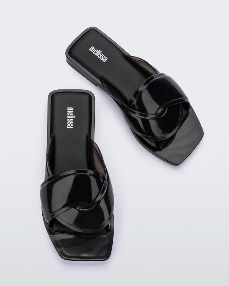 An angled top view of a pair of black Melissa Brigitte slides with a weft designed upper strap.
