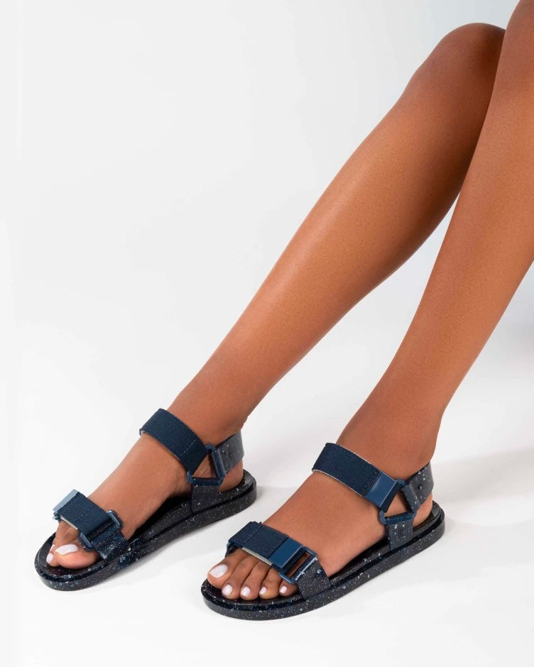 Model's legs wearing a pair of blue/silver glitter Melissa Wide Papete sandals with velcro ankle and front straps.