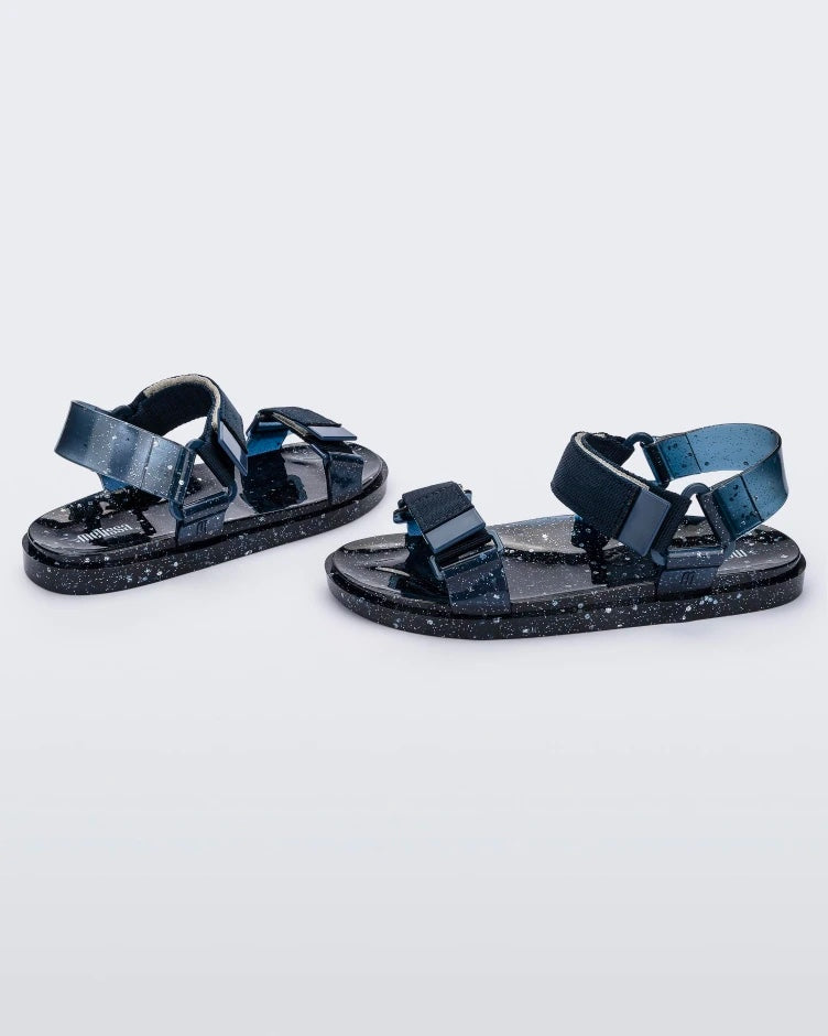Angled view of a pair of blue/silver glitter Melissa Wide Papete sandals with velcro ankle and front straps.
