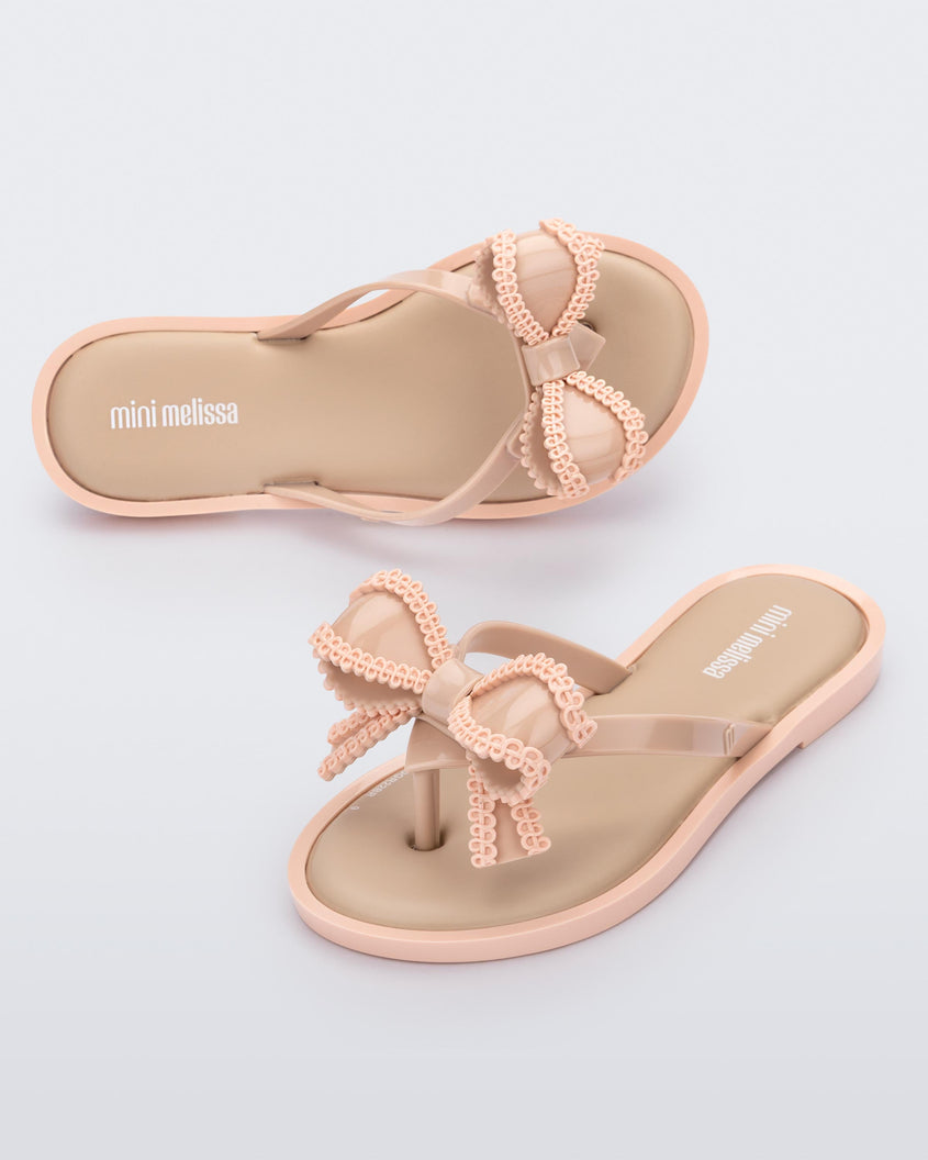 An angled front and top view of a pair of beige Mini Melissa Slim flip flops with a lace like bow detail.