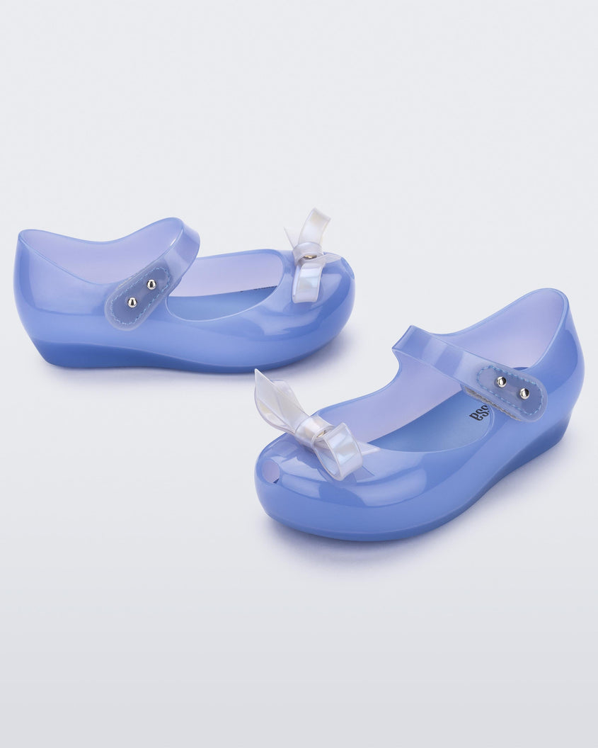 An angled front and side view of a pair of Blue Mini Melissa Ultragirl Bow flats with a blue base, top strap and white bow detail on the front.