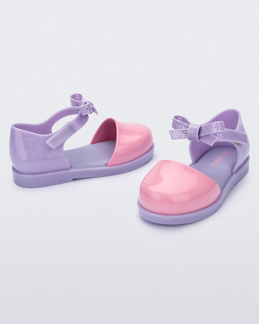 An angled front and side view of a pair of Lilac/Pink Mini Melissa Amy sandals with a pink closed toe section and a back lilac strap with a lace like bow detail.