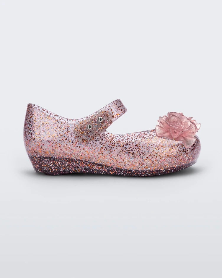 Side view of the Mini Melissa Ultragirl ballet flatin Clear Glitter Pink colorway with 3D printed flower applique on the top of the toe. There is a single strap across the top of the foot with a double snap closure.