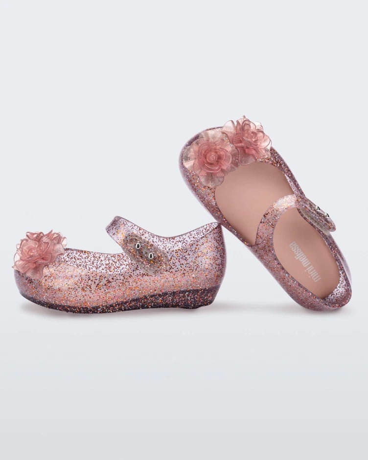 Top and inner side view of a pair of Mini Melissa Ultragirl ballet flats in Clear Glitter Pink colorway with 3D printed flower appliques on the top of the toe. There is a single strap across the top of the foot with a double snap closure.